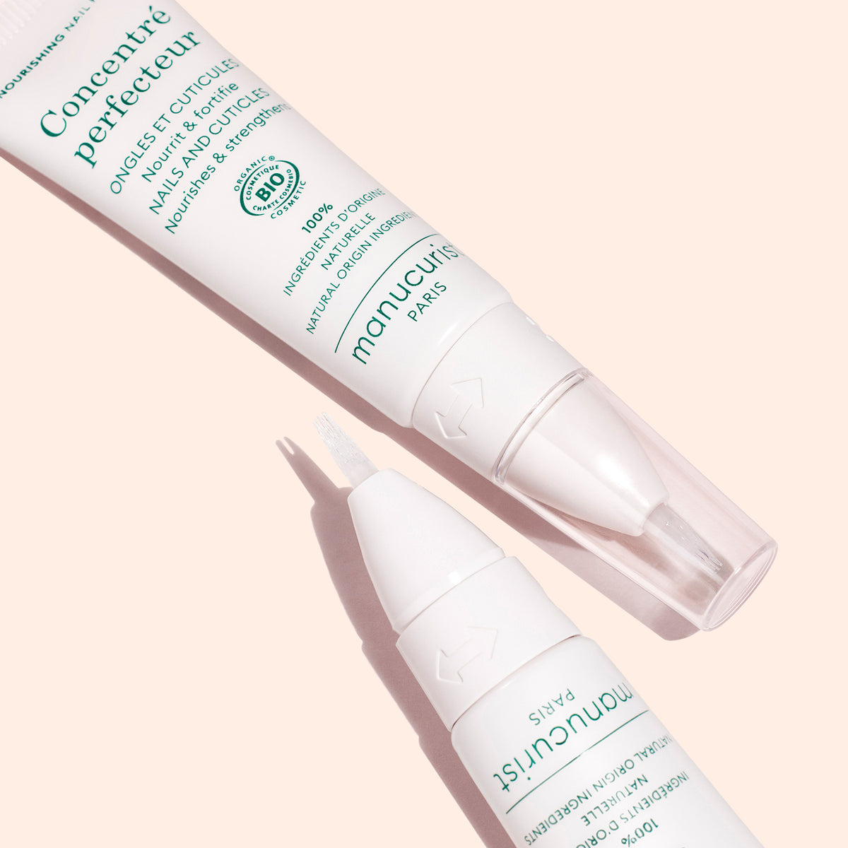 Deux stylos de manucure blancs du Stronger Nails Duo de Manucurist, présentés sur un fond pêche. L’un est fermé et l’autre révèle un embout pinceau. Tous deux arborent un texte vert et portent la mention « Concentré perfecteur ».