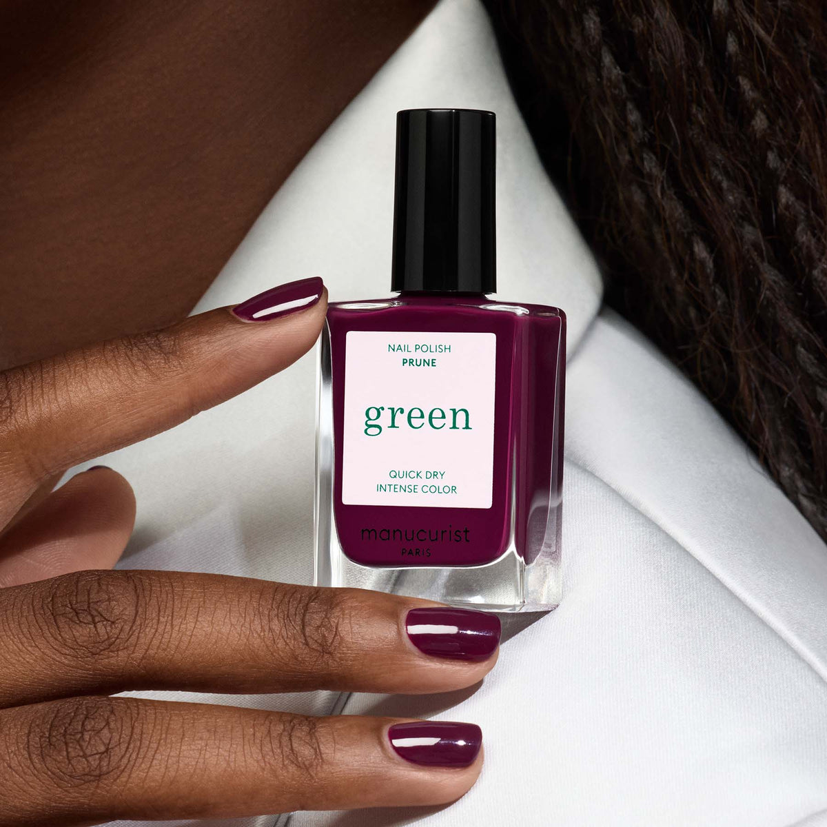 A hand with dark skin tone shows neatly manicured nails painted in Manucurist Prune, a deep purple quick-dry polish, holding a matching bottle against a white background.
