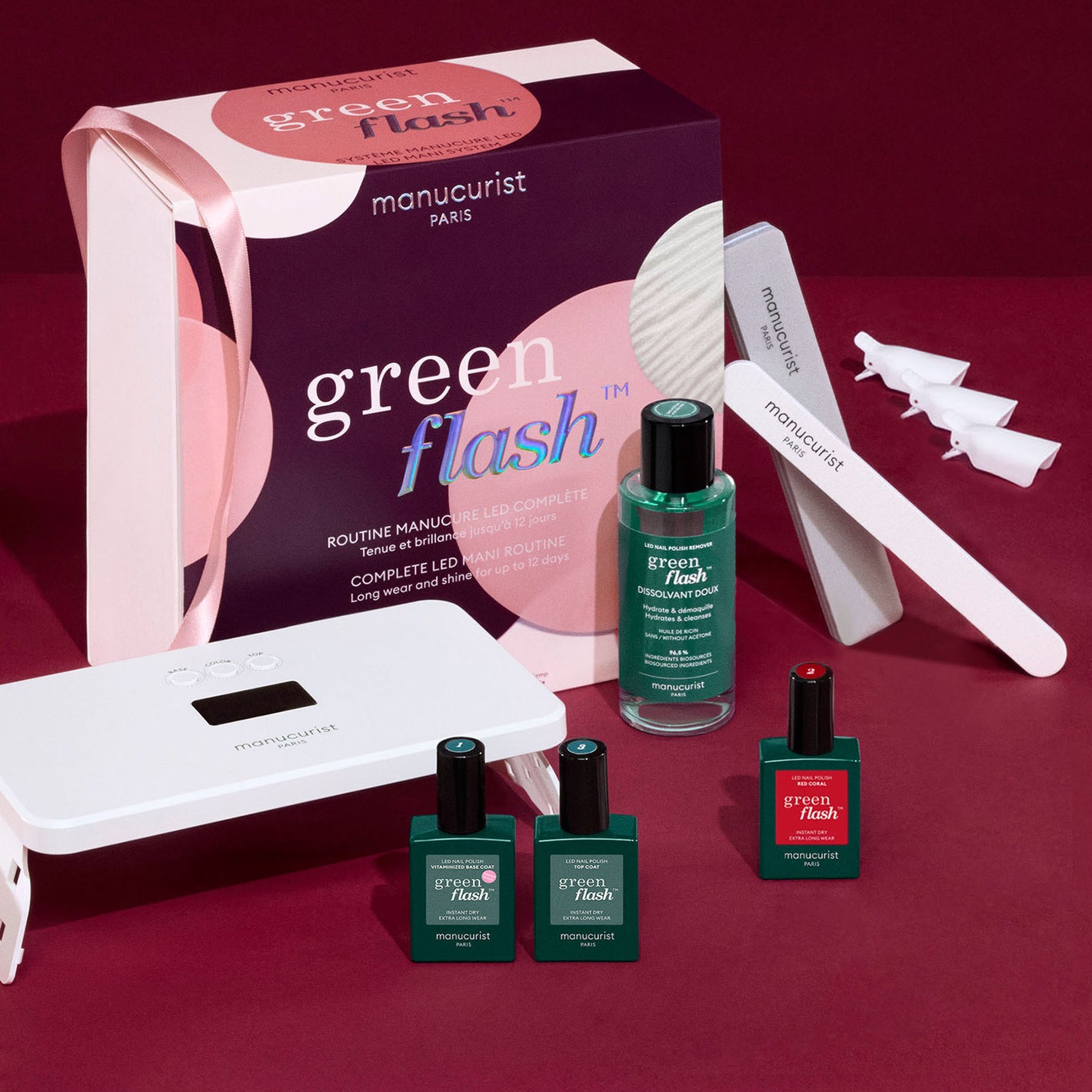 A Manucurist Green Flash manicure kit is displayed on a red surface, including a boxed set, two nail polish bottles, a remover, LED lamp, nail file, four clips, and a cuticle stick.