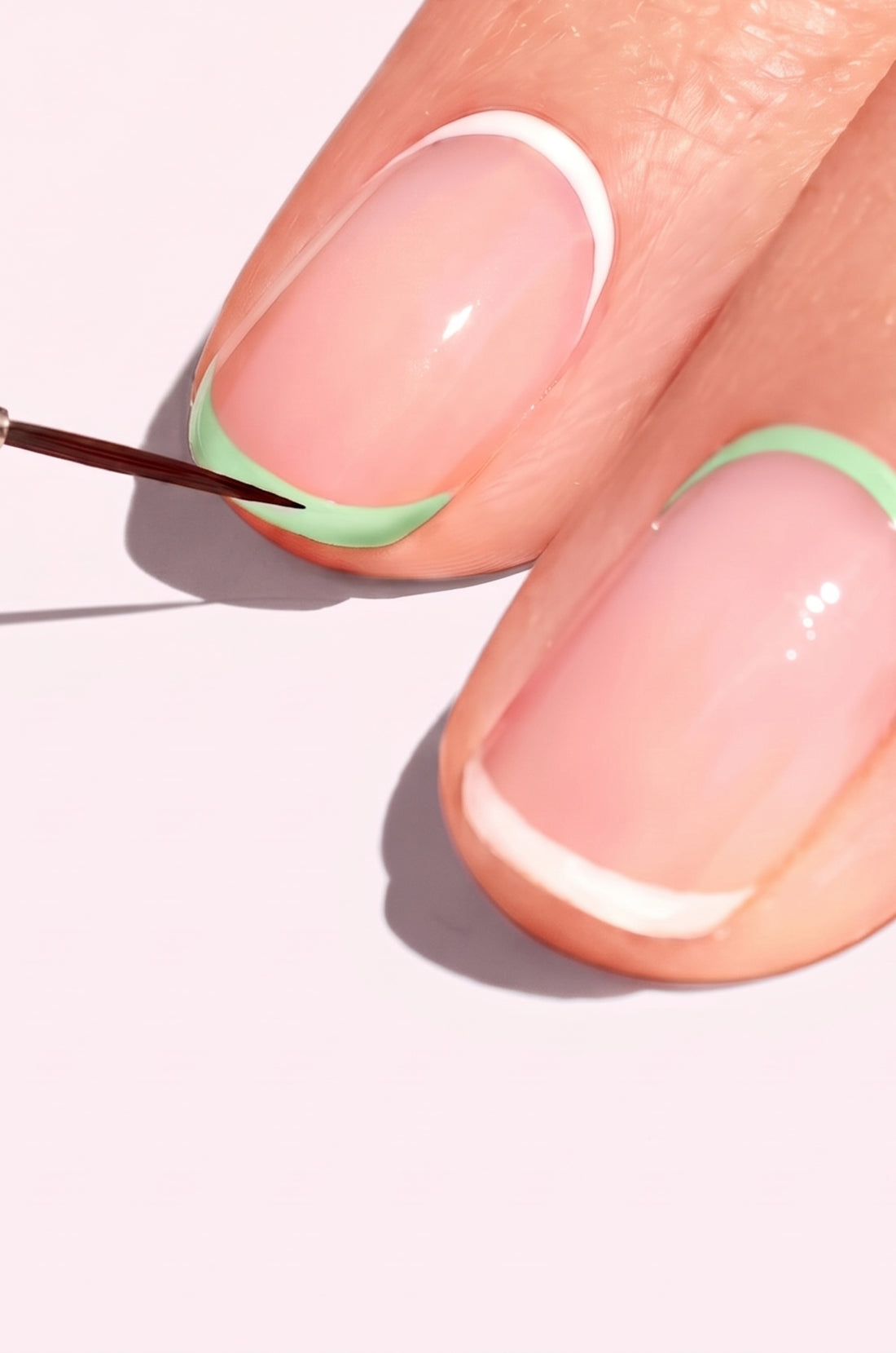 A close-up of fingernails with a French manicure; one nail tip has a green line being painted while others have white tips, all on a pale background.