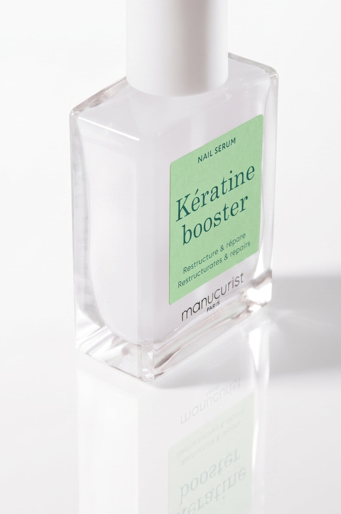 A clear glass bottle of Manucurist Paris Kératine Booster nail serum with a white cap, standing on a white reflective surface. The label is green with dark green text: NAIL SERUM, Kératine booster, and Restructure  répare.