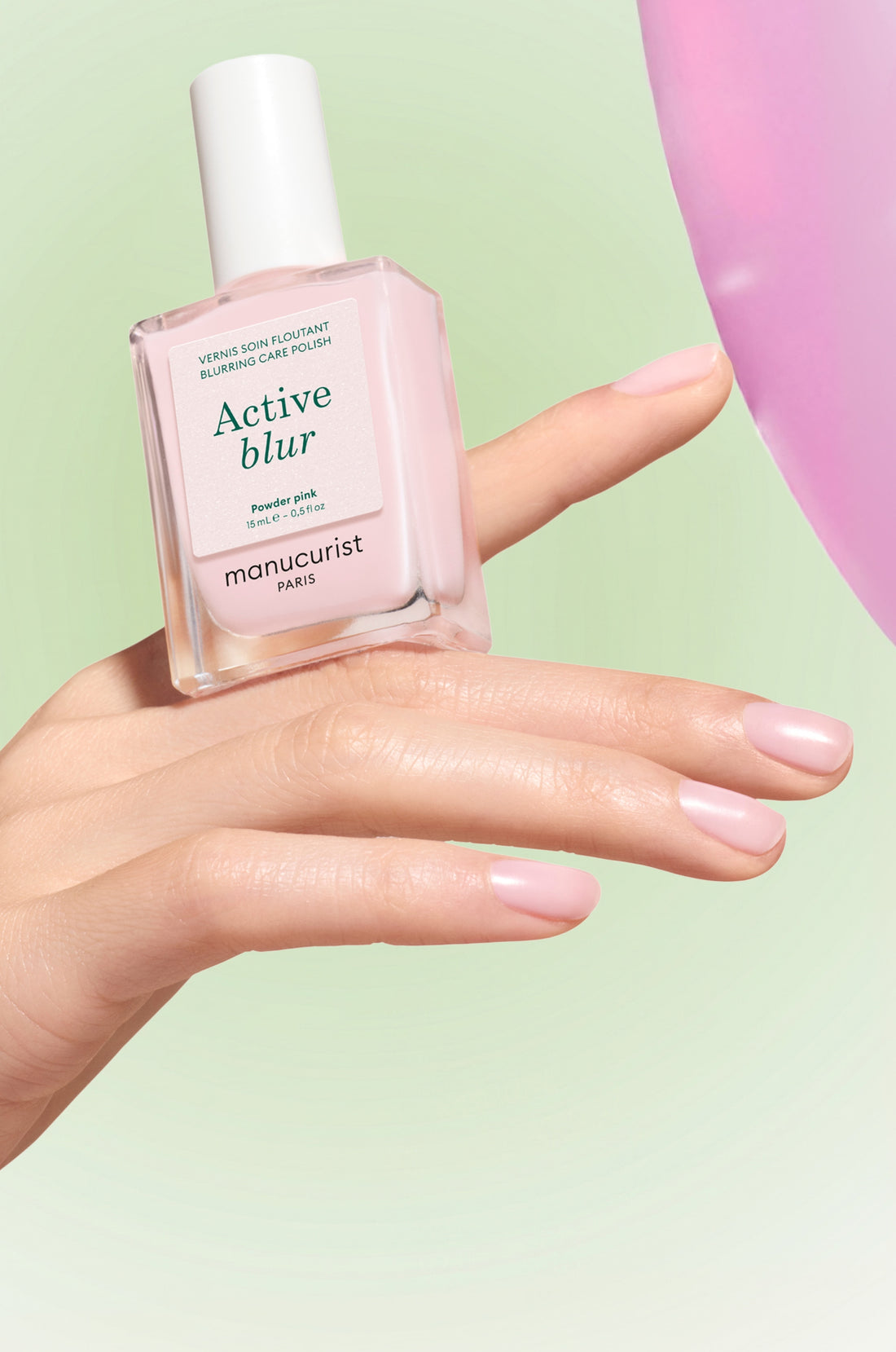 A hand with manicured nails holds a bottle of Manicurist Paris Active Blur blurring care polish against a soft green background, with a pink balloon on the right.