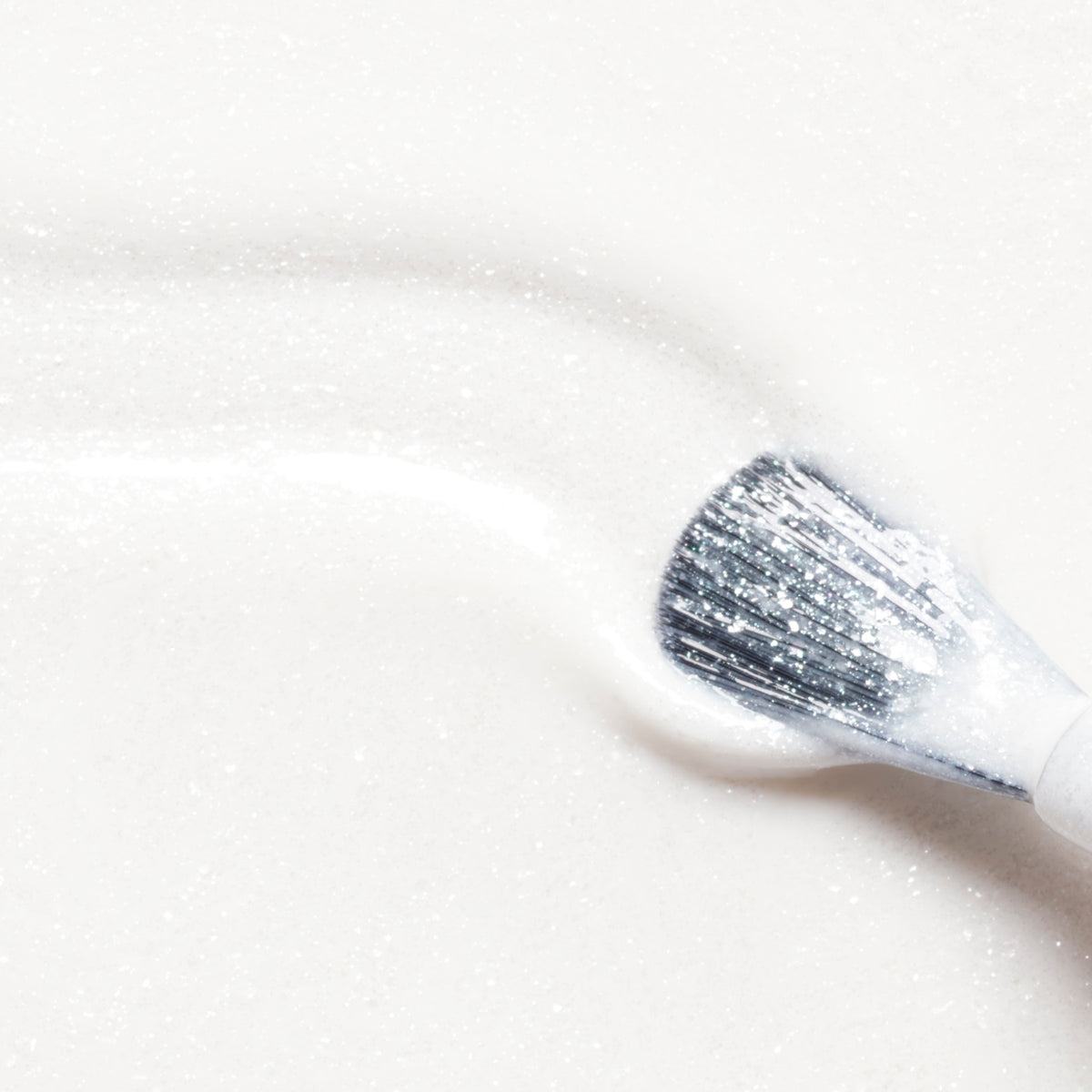 A makeup brush sweeps Manucurist’s Bliss across a smooth, sparkling surface, leaving a visible trail of iridescent white radiance reminiscent of the Green Flash or the glossy brilliance of gel polish.