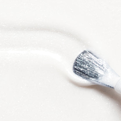 A makeup brush sweeps Manucurist’s Bliss across a smooth, sparkling surface, leaving a visible trail of iridescent white radiance reminiscent of the Green Flash or the glossy brilliance of gel polish.