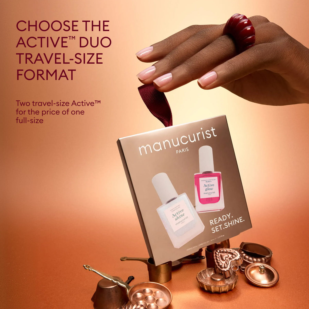 A hand with a ribbon holds a box of Manucurist Active™ Shine Icy travel-size nail polish, perfect for on-the-go nail care. The background features small decorative cups and a bronze teapot on a brown surface. Text highlights the travel-size duo offer.