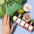 Une main aux ongles multicolores repose sur un tissu rayé à côté de cinq flacons de vernis à ongles Manucurist « Glitter in Love » à étiquette verte, de paillettes biodégradables, de pinceaux fins pour Nail Art et d&