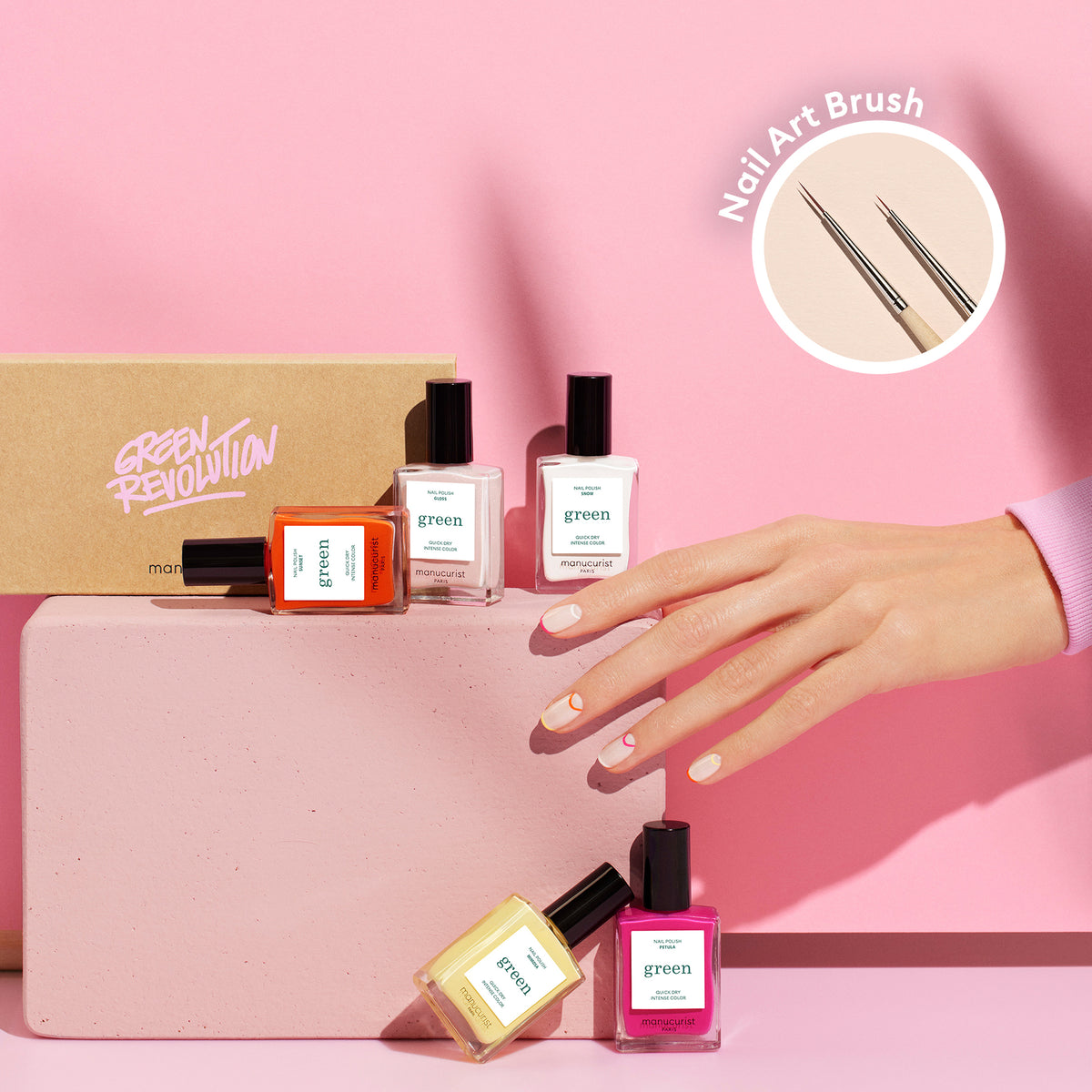 Une main arborant un Nail Art estival pastel de la collection Smile! par Manucurist tient un bloc rose, entourée de quatre flacons de vernis à ongles Green Revolution et d&