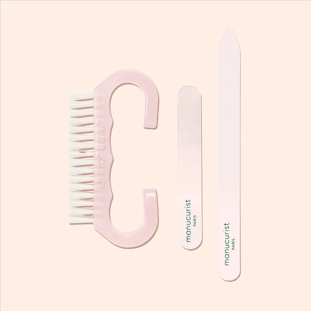 A pink Manucurist-us Nail Brush with vegan bristles, accompanied by a compact emery board and a long white nail file featuring the manucurist PARIS logo—all ideal for an at-home manicure—are displayed neatly on a light beige background.
