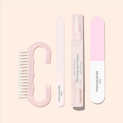 A Manucurist-us Nail Brush featuring vegan bristles in pink, accompanied by a white nail file, a clear Peeling Glycolique tube, and a pink and white nail buffer—all essential tools for the perfect manicure—are arranged neatly on a beige background.