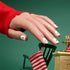 A hand adorned with Manucurist Stickers Nail Art Holidays—white polish decorated with red stars and crescent moons—peeks out from a red sweater, hovering above a small green chair and a gold ornament wearing a striped red and white stocking, offering perfect festive manicure inspiration.