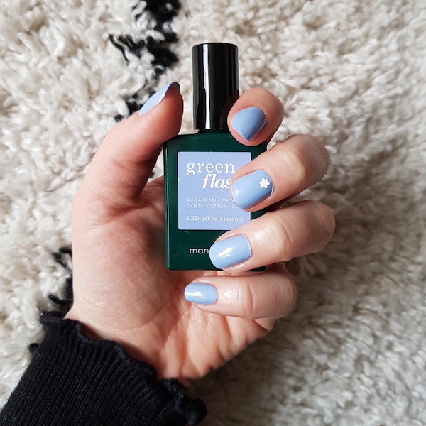 Une main aux ongles peints en bleu clair, dont l’un arbore un petit motif de fleur blanche, tient un flacon de vernis à ongles vert étiqueté Green Flash™. L’arrière-plan est un tissu texturé crème et noir.
