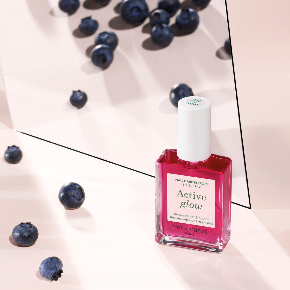 Un flacon de vernis soin Manucurist Active™ Glow Blueberry repose sur une surface rose entourée de myrtilles éparpillées, avec un miroir réfléchissant montrant l’arrière du flacon.