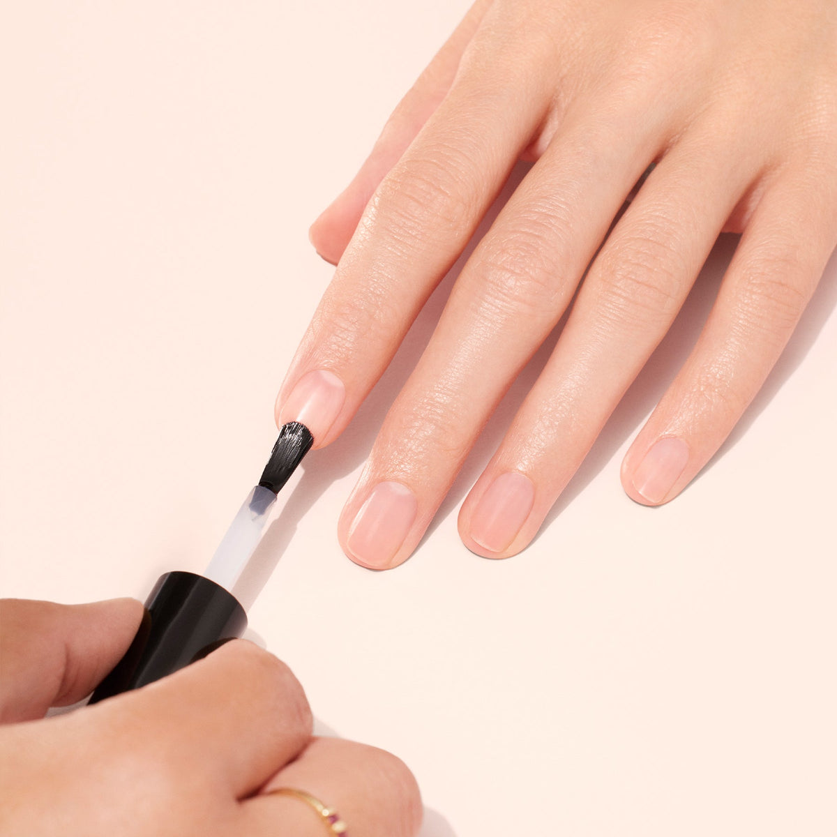 Une main aux ongles soigneusement taillés reçoit une application de vernis à ongles transparent à l'aide d'un pinceau, posée sur une surface de couleur claire.