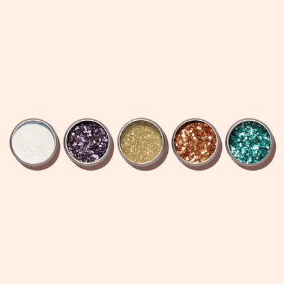 Five small round containers of Manucurist’s Lilac Biodegradable Glitter, each in white, purple, gold, copper, and teal—perfect for nail art—are arranged in a row on a light beige background.