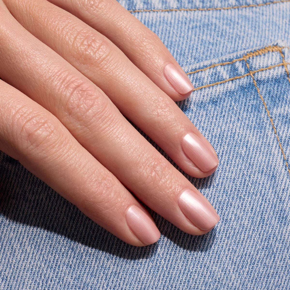 Une main aux ongles soigneusement manucurés, vernis avec le gel Carnation de Manucurist, repose sur un tissu en denim bleu clair.