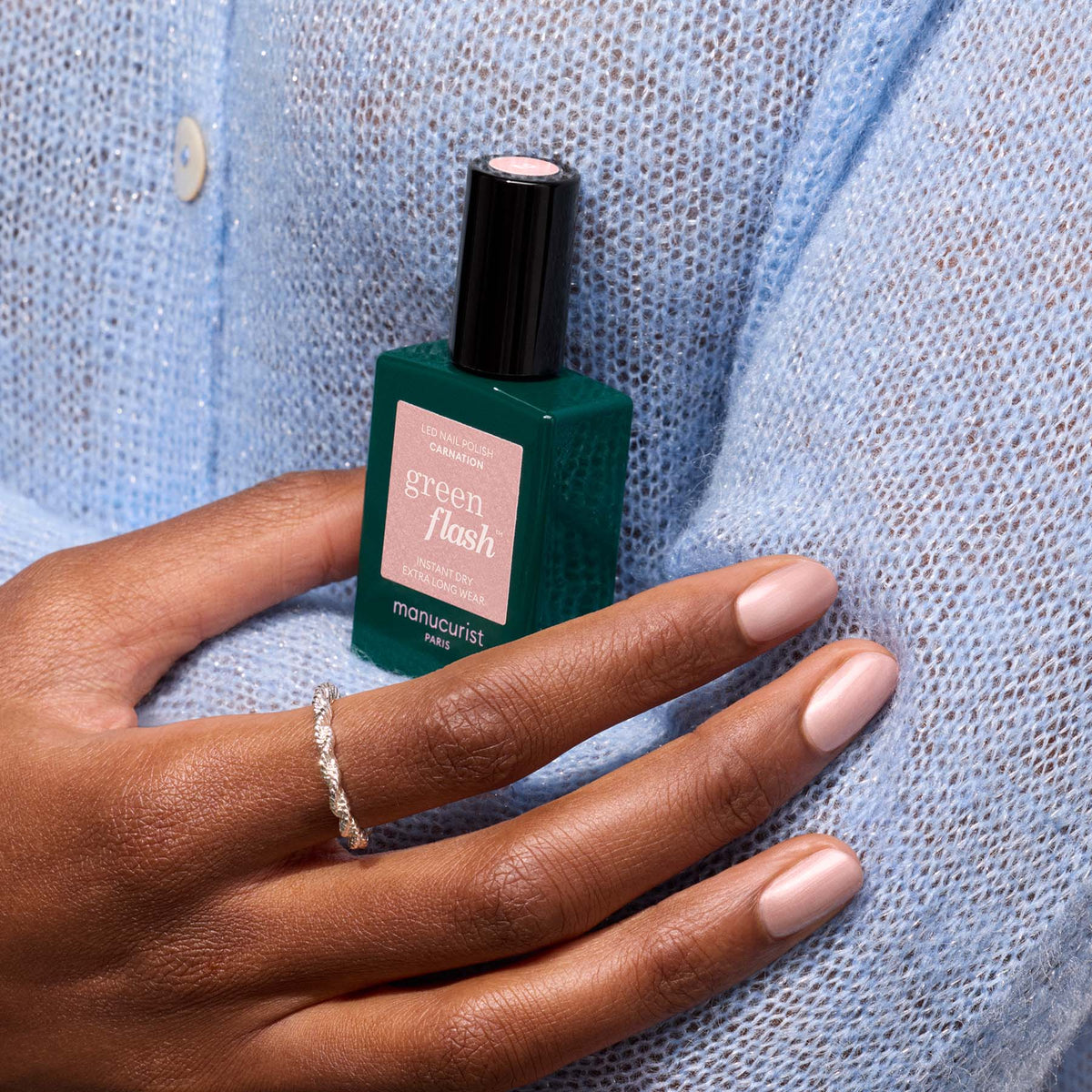 Une personne portant un pull en maille bleu clair tient un flacon de vernis gel Carnation de Manucurist. Ses ongles sont manucurés dans une teinte nude rose nacré, sublimée par une délicate bague en argent à son doigt.