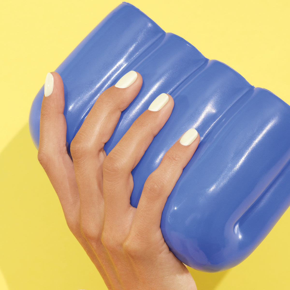 A hand with neatly manicured, creamy Porcelaine polish by Manucurist holds a glossy, blue, rounded object against a bright yellow background.