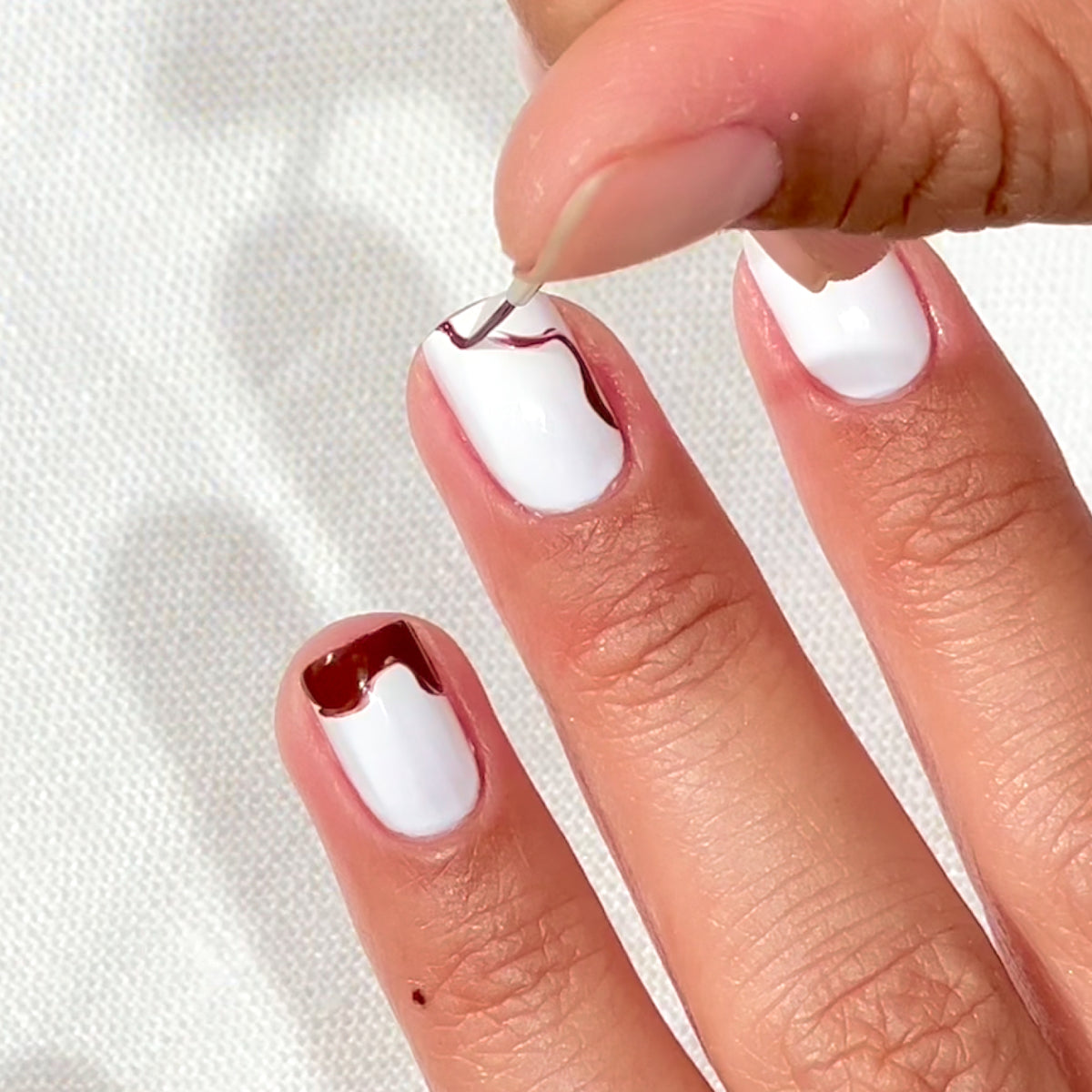 Gros plan sur une main aux ongles vernis de blanc. Une personne utilise un pinceau fin pour dessiner un motif ondulé marron sur le bout et le côté d'un ongle, sur un fond blanc texturé.