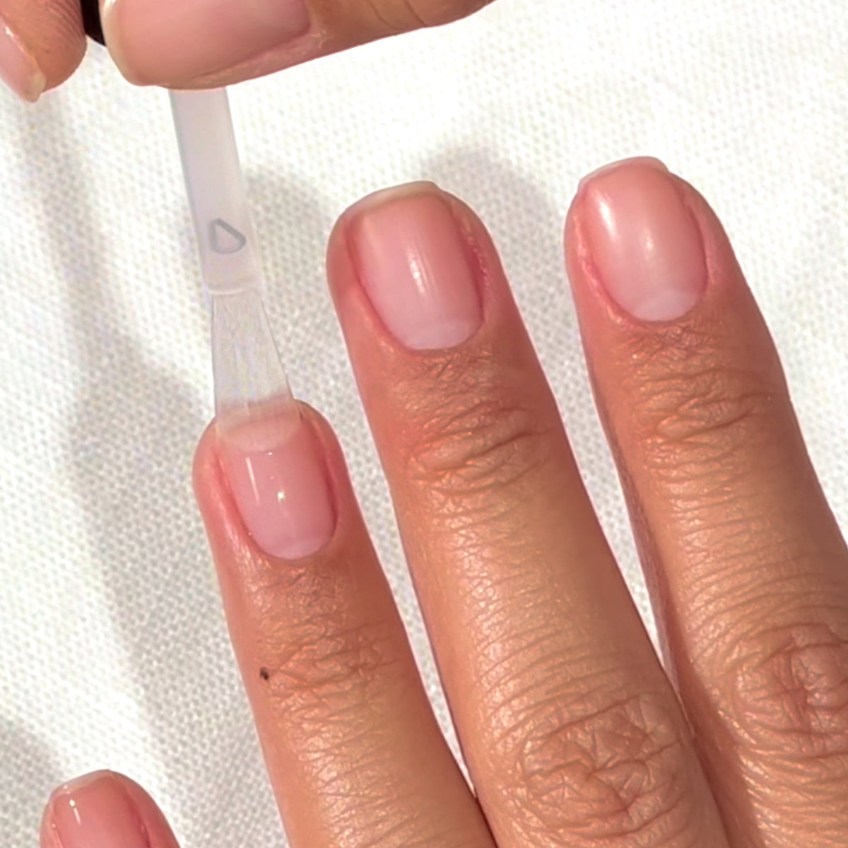Une main aux ongles propres et naturels est présentée pendant l’application d’un vernis à ongles transparent sur l’ongle du pouce à l’aide d’un pinceau. Le fond est une surface blanche texturée.