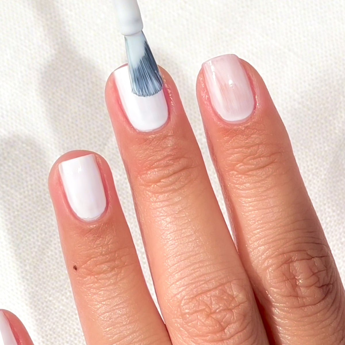 Gros plan sur une main laissant apparaître trois doigts, dont les ongles sont vernis de blanc à l'aide d'un pinceau, sur un fond blanc texturé.