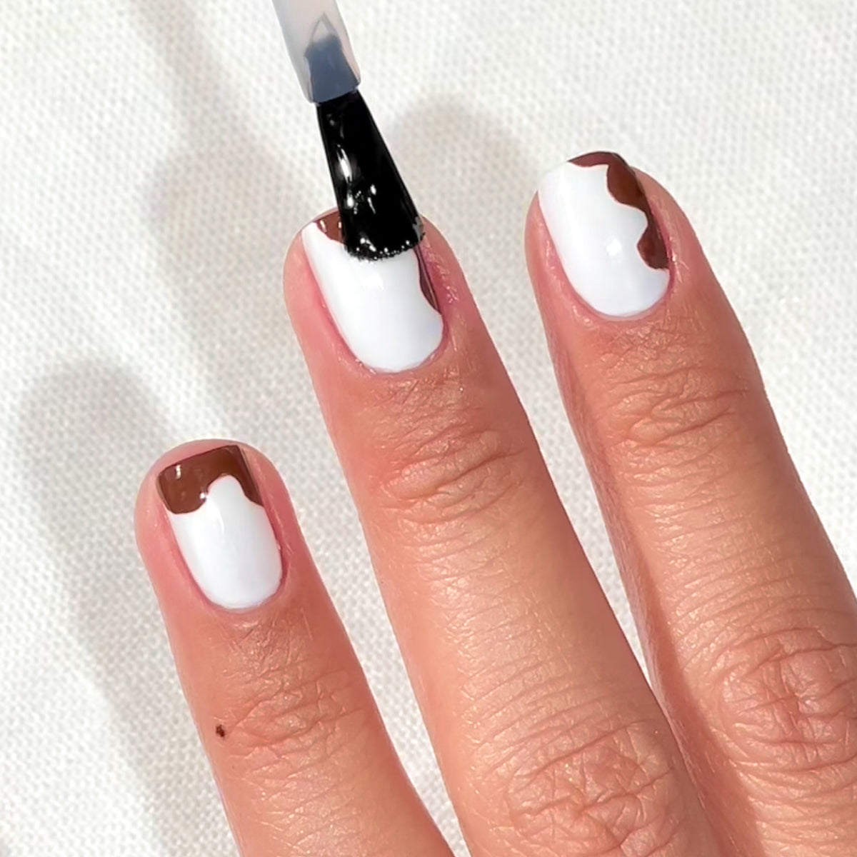 Une main avec du vernis blanc et des motifs abstraits marron sur des ongles courts, avec un pinceau appliquant un Top Coat transparent sur un ongle. L’arrière-plan est une surface blanche texturée.