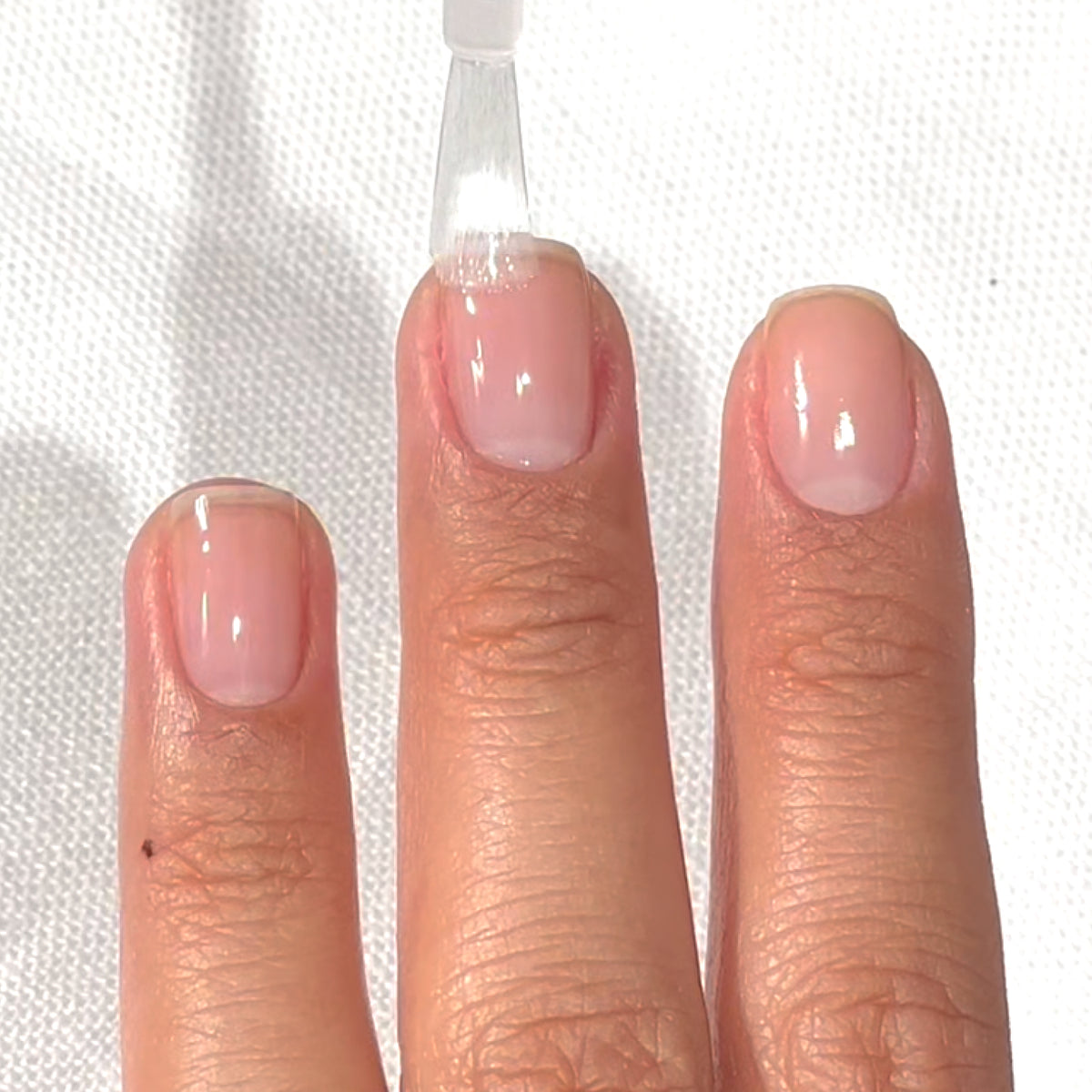 Une main aux ongles courts et naturels est montrée tandis qu'un vernis transparent est appliqué sur l'ongle du majeur, au-dessus d'une surface claire.