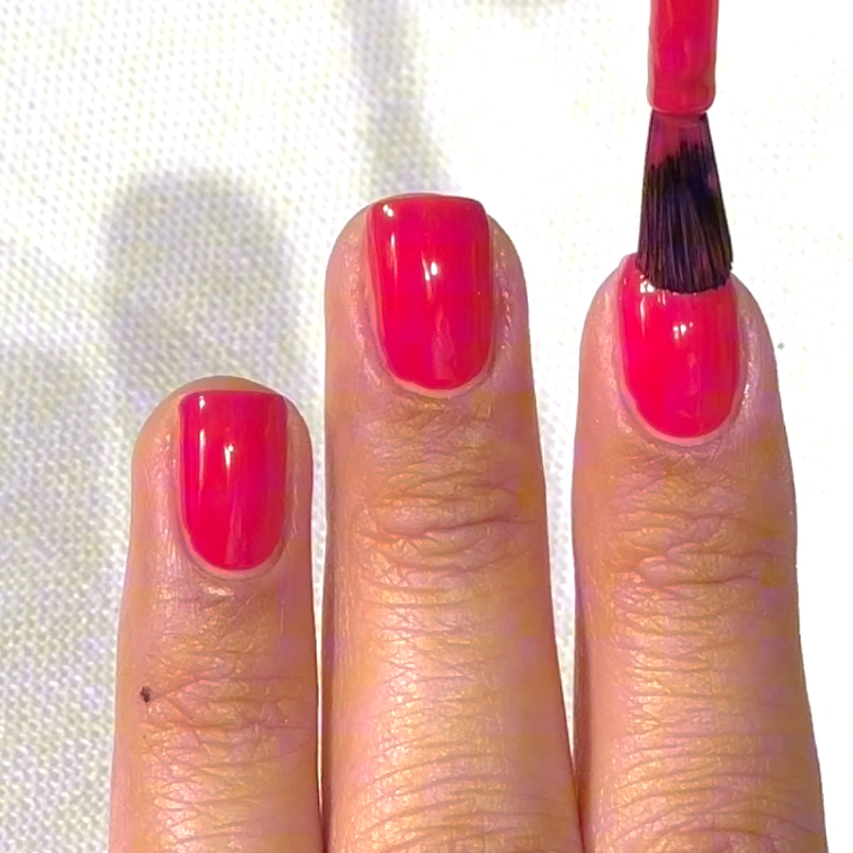 Une main aux ongles courts en train de se faire appliquer un vernis à ongles rose vif sur le majeur à l'aide d'un pinceau. Les autres ongles sont déjà vernis.