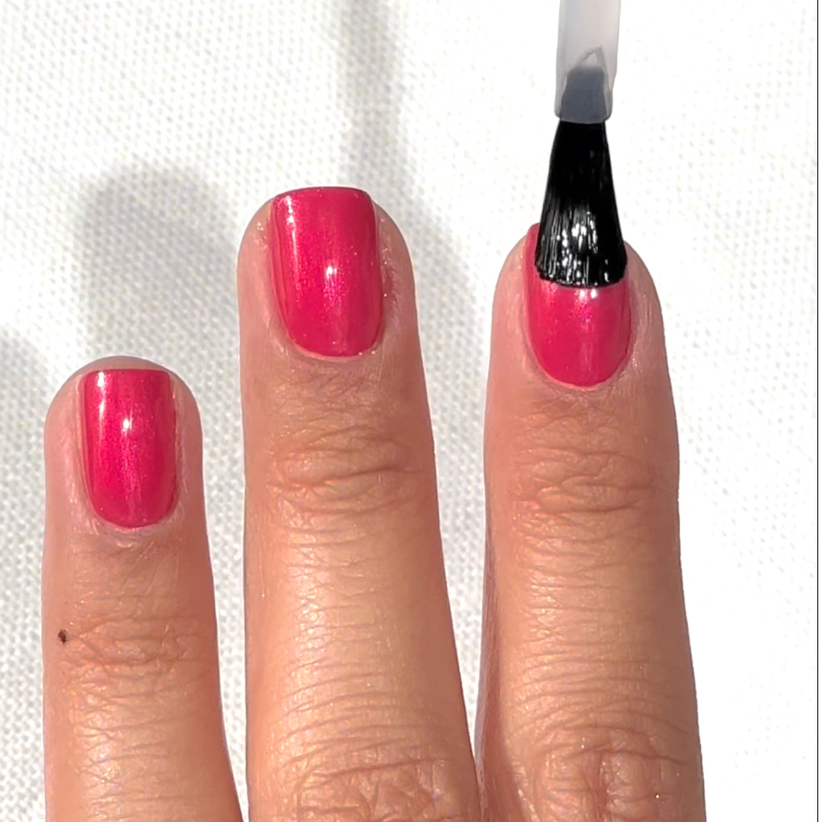 Une main aux ongles vernis roses est présentée, tandis qu’un pinceau applique un Top Coat transparent sur l’ongle du majeur. L’arrière-plan est une surface blanche texturée.