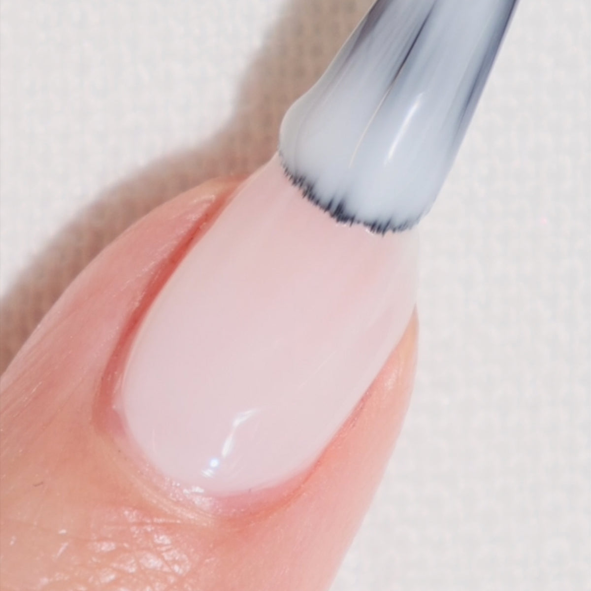 A close-up of a fingernail being painted with light pink nail polish using a brush, against a white textured background.