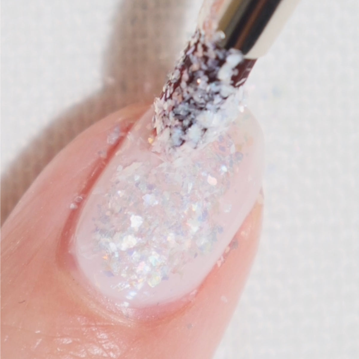 A close-up of a fingernail being painted with a brush applying a layer of translucent glittery nail polish, featuring white and iridescent sparkles.