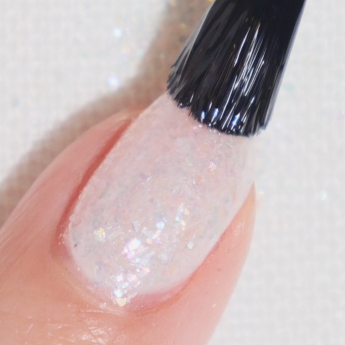 A close-up of a fingernail being painted with a shimmery, iridescent white nail polish using a black-bristled brush.