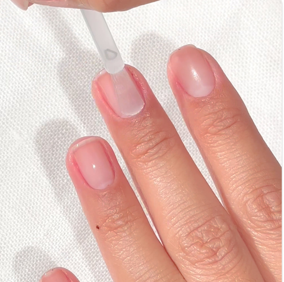 A close-up of a hand with short, natural nails as clear nail polish is being applied to the fingernail of the middle finger using a brush. The background is a white textured surface.