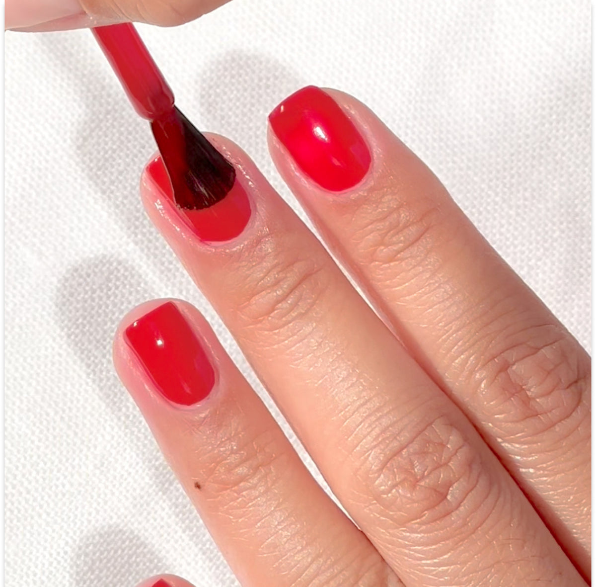 A hand with neatly trimmed nails is having bright red nail polish applied to the fingernails using a brush, with a white textured background underneath.