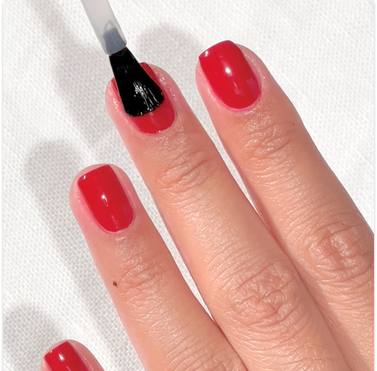 A hand with red-painted nails is shown, and a brush applies a clear top coat to the middle fingernail. The background is a white textured surface.