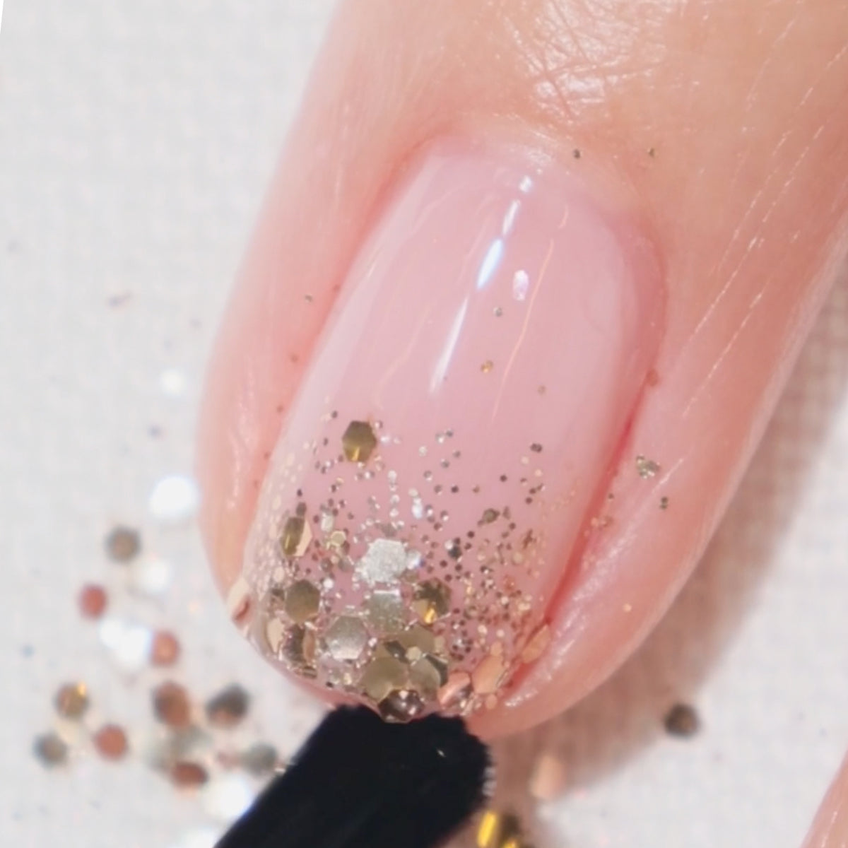 Gros plan sur un ongle avec une base rose clair et des paillettes dorées appliquées près du bord libre, finalisé avec un Top Coat transparent. Des paillettes libres sont visibles sur la surface autour.