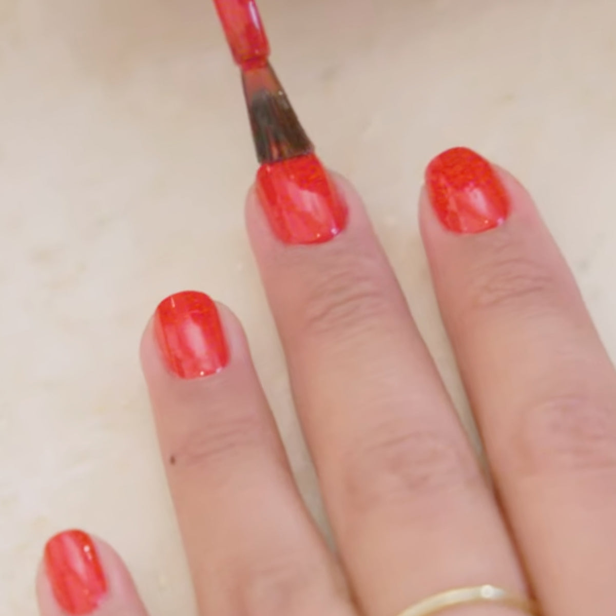 A close-up of a hand with a gold ring on one finger, having red nail polish applied to the nails with a brush.