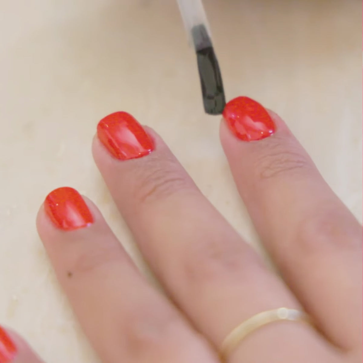 A hand with red-painted nails is having a clear top coat applied. The person is wearing a thin gold ring on their finger.