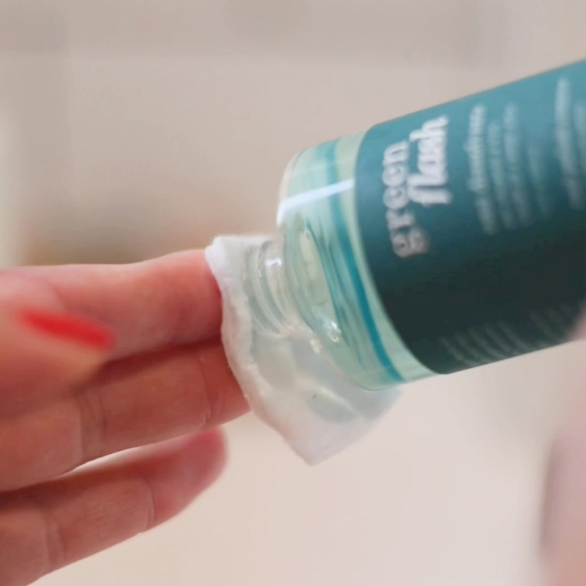 A hand holds a cotton pad while pouring a clear liquid from a bottle labeled Green Flash onto the pad. The focus is on the action, with the background blurred.