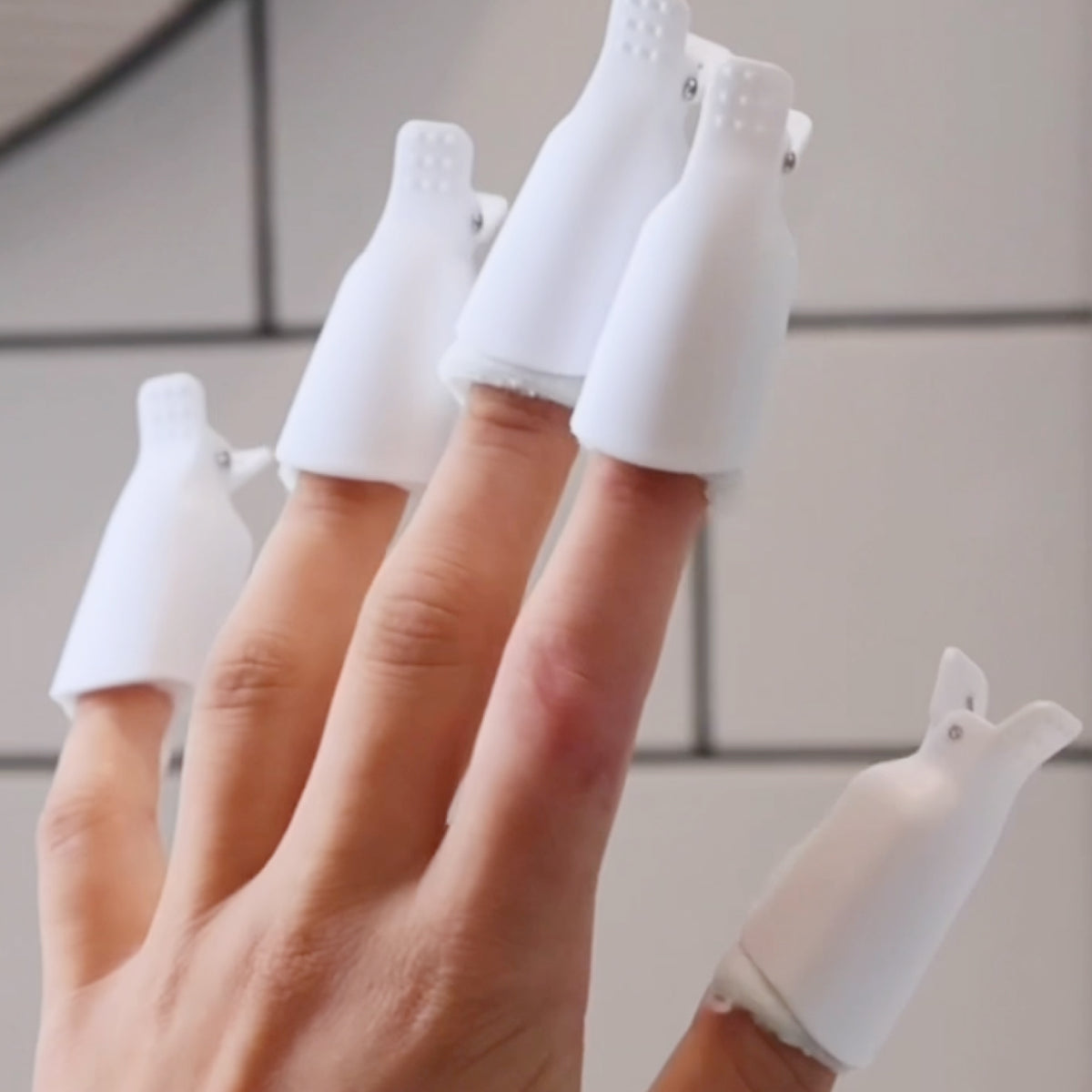 A hand with finger covers shaped like small white bottles or clips on each finger, set against a tiled background.