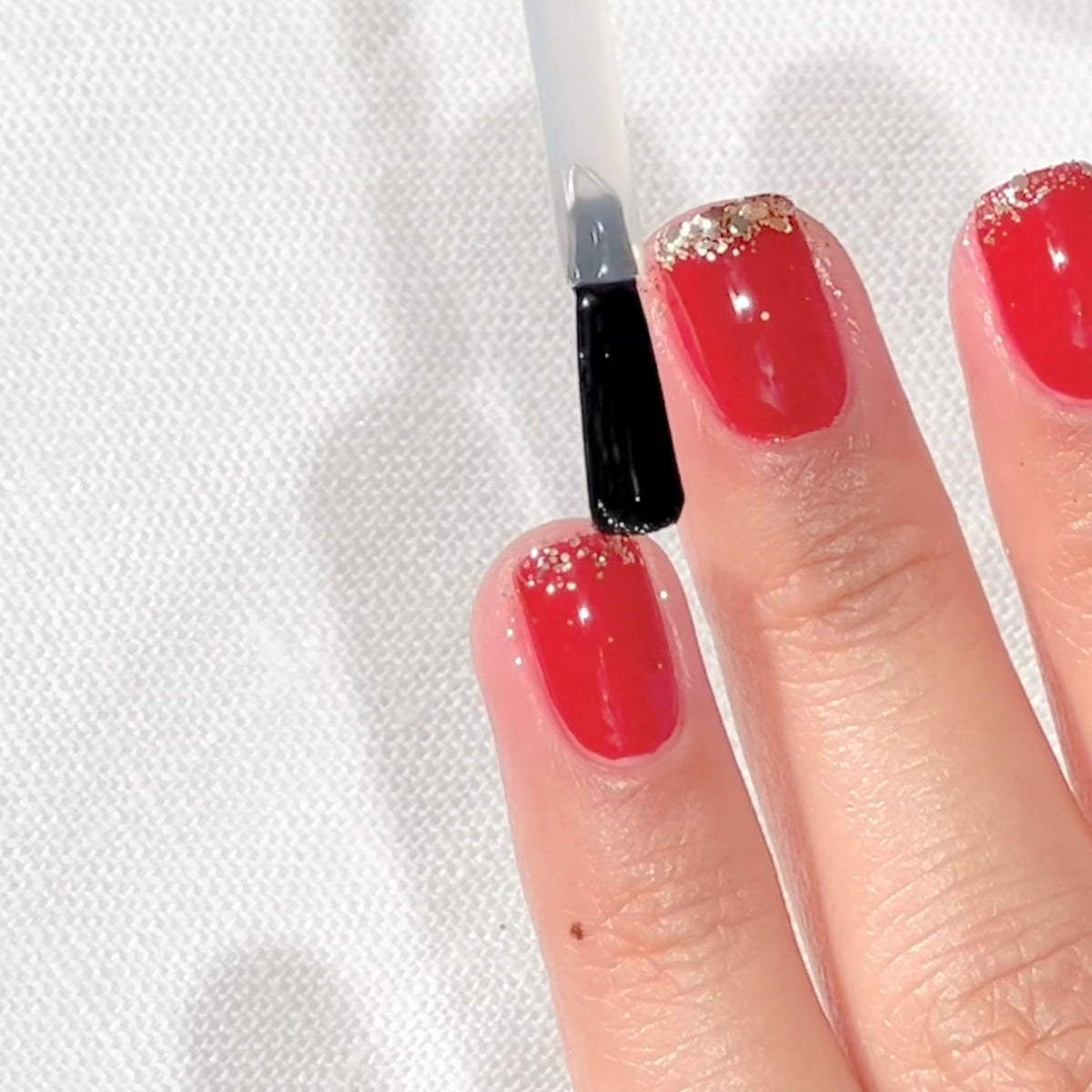 A hand with red nail polish and gold glitter tips is having a clear top coat applied to the middle fingernail using a brush.