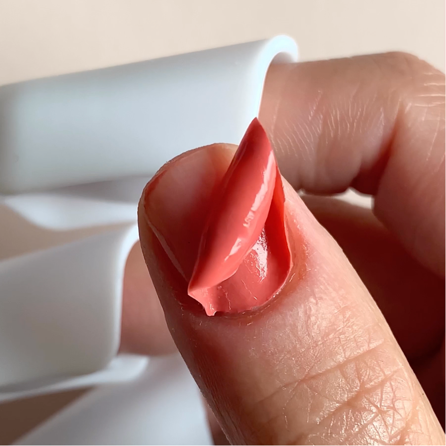 A close-up of a thumb with dried, reddish-pink nail polish peeling off in one piece, being lifted by the persons other hand. The background is out of focus and light-colored.