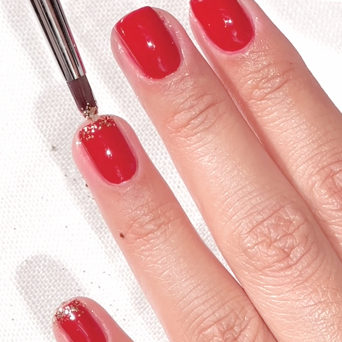 A hand with red nail polish is being decorated with gold glitter at the base of the nails using a small brush.