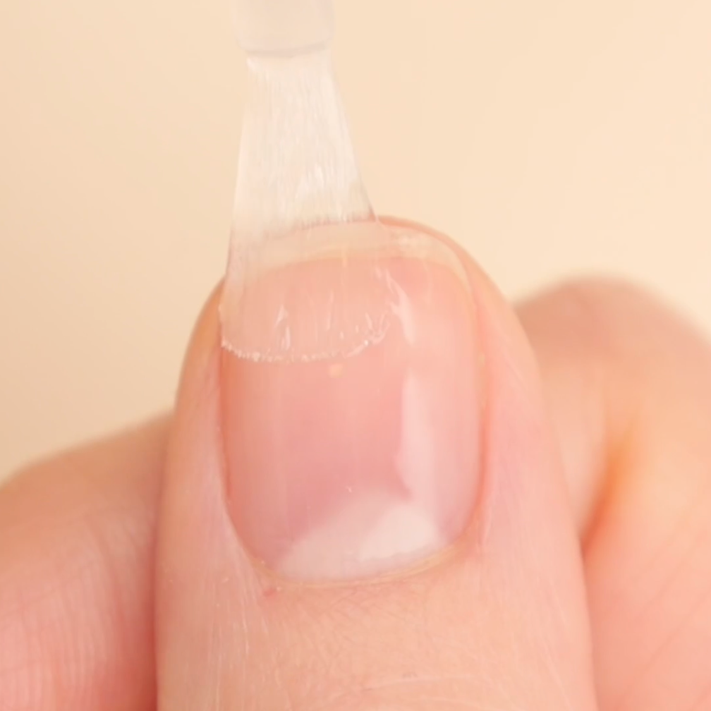 A close-up of a fingernail with a brush applying a clear coat of nail polish. The nail and surrounding skin are clean and well-groomed, and the background is a soft beige color.