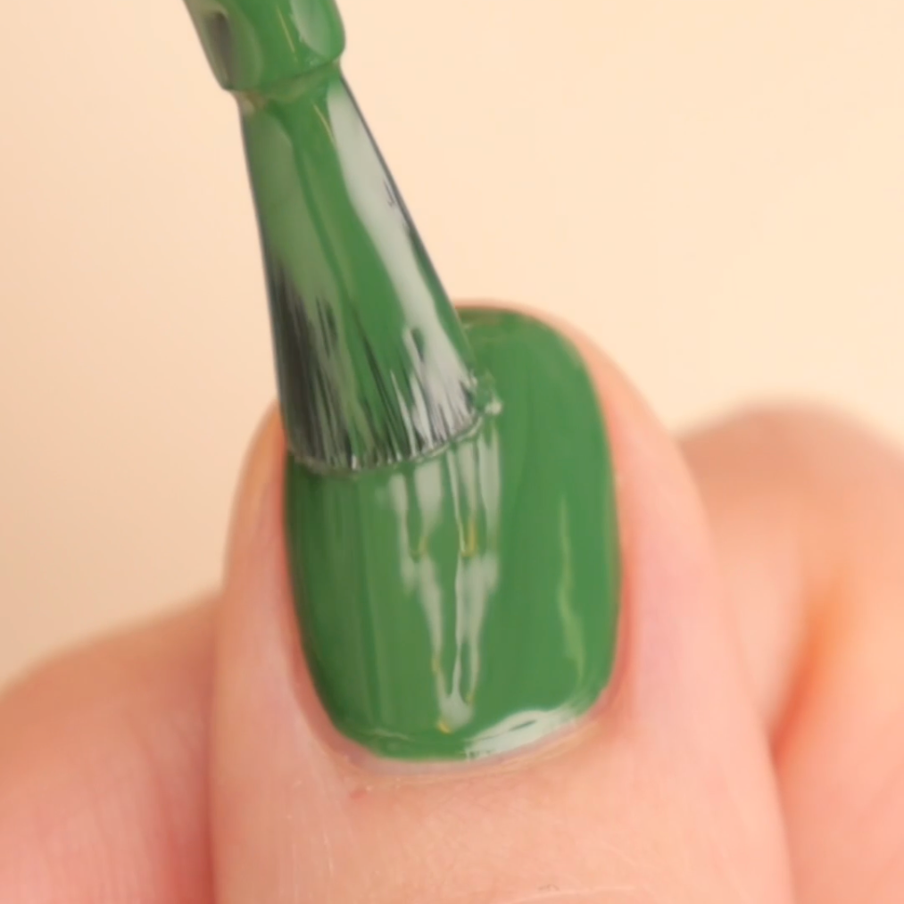 A close-up of a fingernail being painted with glossy green nail polish using a brush.