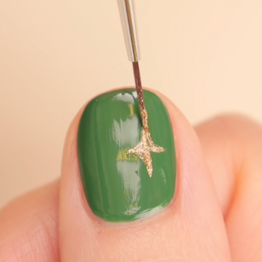 A close-up of a fingernail painted green, with a fine brush applying a gold, star-like design on top.