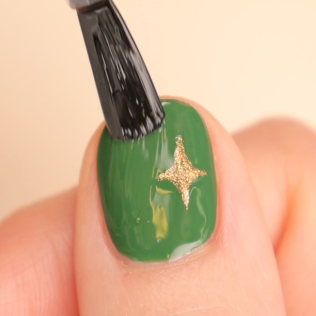 A close-up of a fingernail painted green with a gold star design, as a brush applies a clear top coat over the nail.