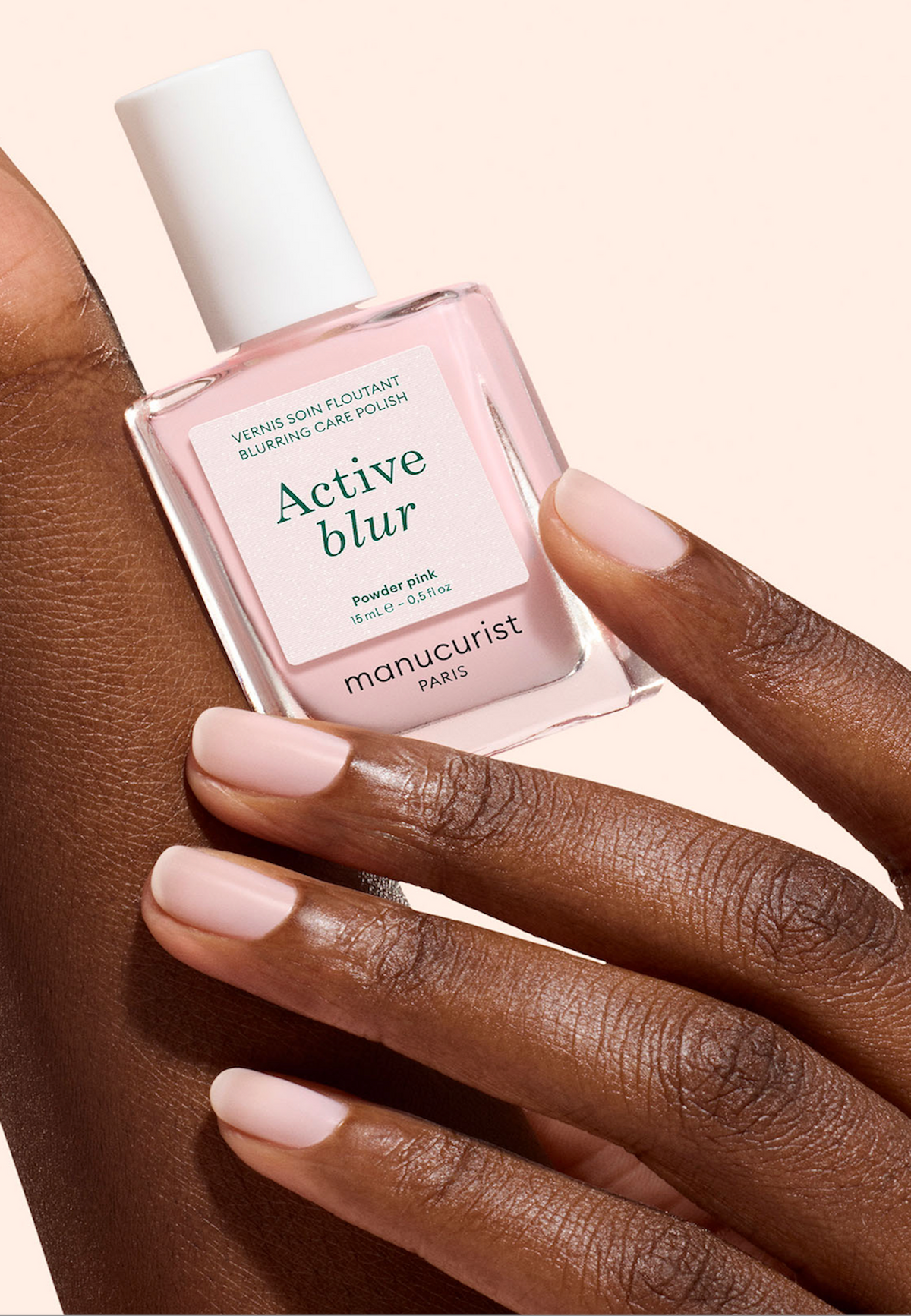 Une main aux ongles soigneusement manucurés, peints dans une teinte rose clair, tient un flacon de vernis à ongles Manucurist Paris Active Blur en Powder Pink sur un fond pâle.