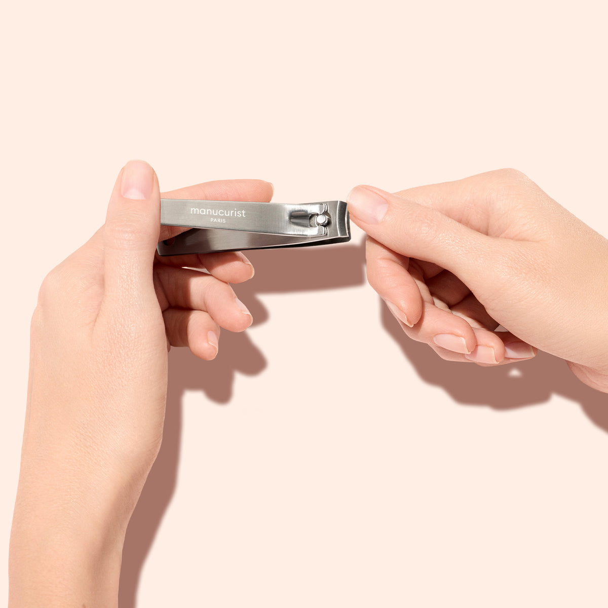 Two hands holding a silver nail clipper against a light beige background. The clipper is labeled manucurist PARIS, and one hand is ready to use it.