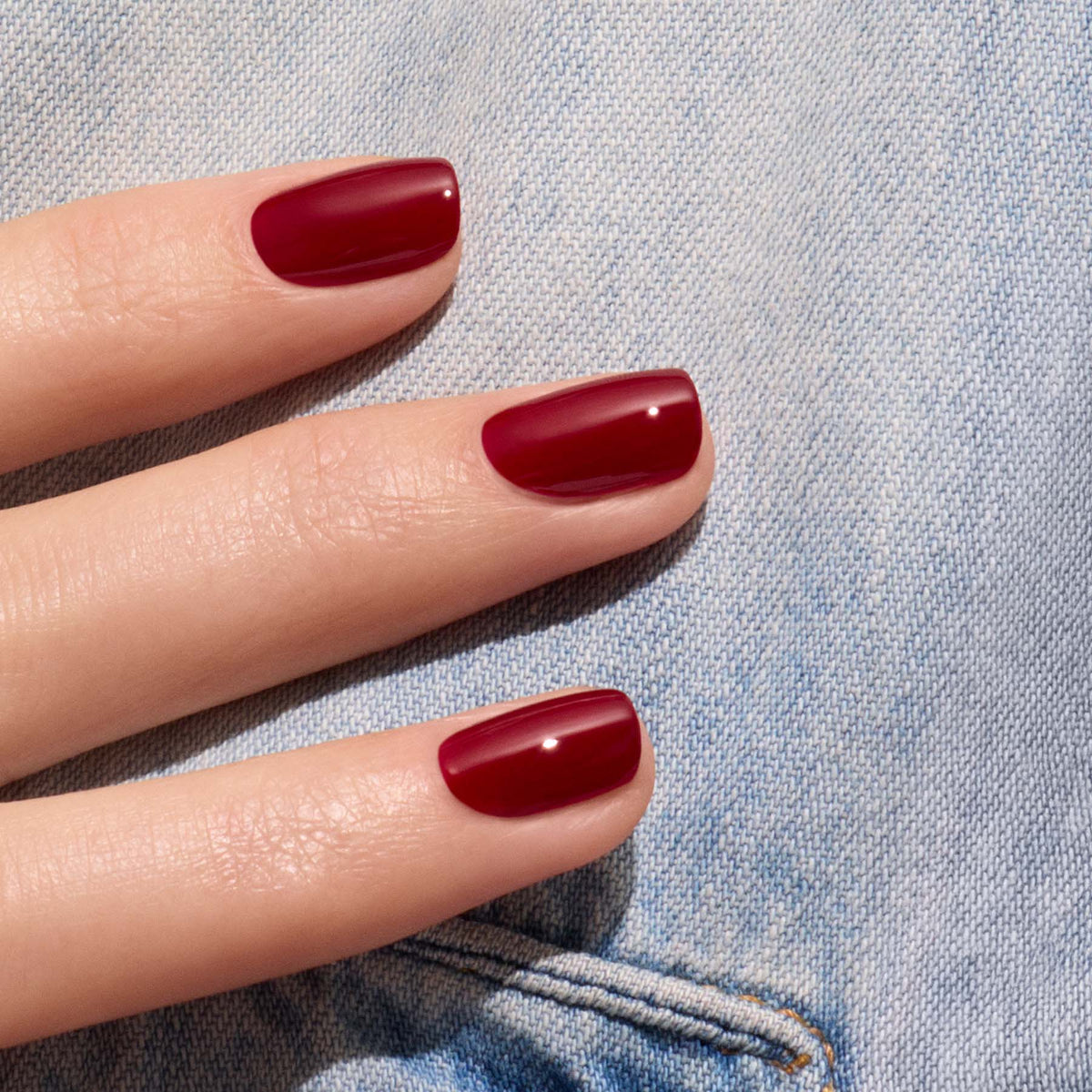Une main aux ongles impeccablement manucurés, vernis dans la teinte rouge velours brillant Dark Pansy de Manucurist, repose sur du denim bleu clair.