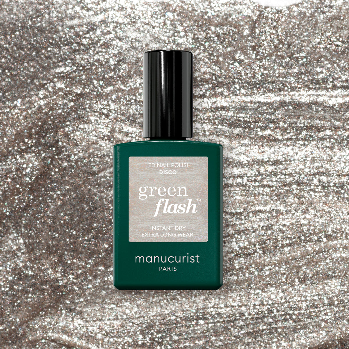 Un flacon de vernis gel Disco de Manucurist repose sur un fond argenté scintillant. Le flacon vert foncé avec un capot noir arbore une étiquette argentée pailletée.